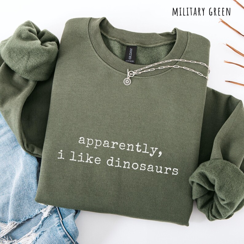 May include: Military green sweatshirt with the text "apparently, i like dinosaurs" in white. A silver necklace with a sunburst pendant is draped over the front. The sweatshirt is folded on top of a pair of blue jeans.