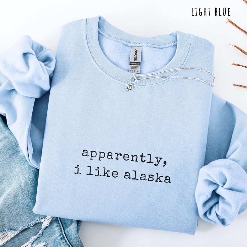 May include: Light blue crewneck sweatshirt with the text "apparently, i like alaska" in black. A silver necklace with a pendant is draped over the sweatshirt. The sweatshirt is folded next to a pair of blue jeans.