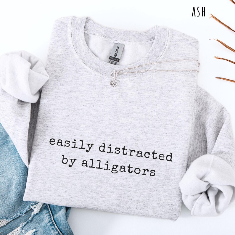 May include: A light gray sweatshirt with the text "easily distracted by alligators" in black. The sweatshirt has long sleeves and a crew neck. A silver necklace with a small pendant is draped across the front. The word "ASH" is in the top right corner.
