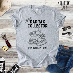 Może przedstawiać: Szara koszulka z napisem "DAD TAX COLLECTOR" i grafiką przedstawiającą jedzenie typu fast food. Koszulka zawiera r&oacute;wnież frazę "IF I'M BUYIN', I'M TRYIN'".