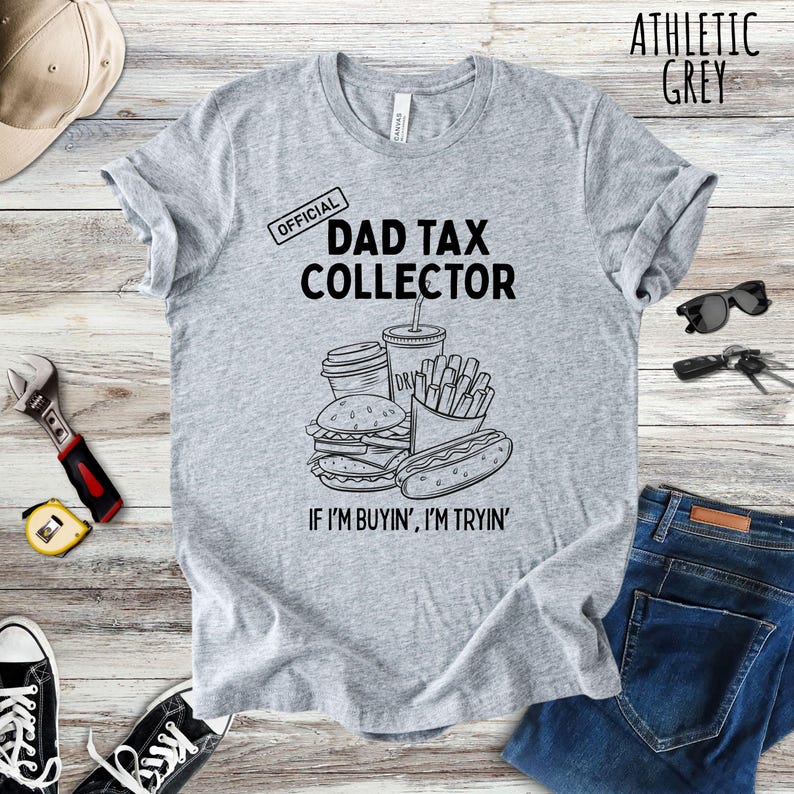 Może przedstawiać: Szara koszulka z napisem "DAD TAX COLLECTOR" i grafiką przedstawiającą jedzenie typu fast food. Koszulka zawiera r&oacute;wnież frazę "IF I'M BUYIN', I'M TRYIN'".