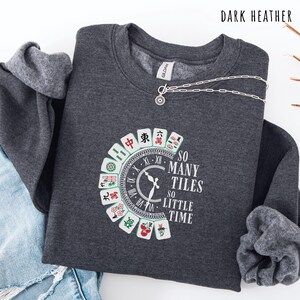 May include: Dark heather gray sweatshirt with a circular design featuring Mahjong tiles and the text "SO MANY TILES SO LITTLE TIME." A silver necklace with a sunburst pendant is draped across the sweatshirt.