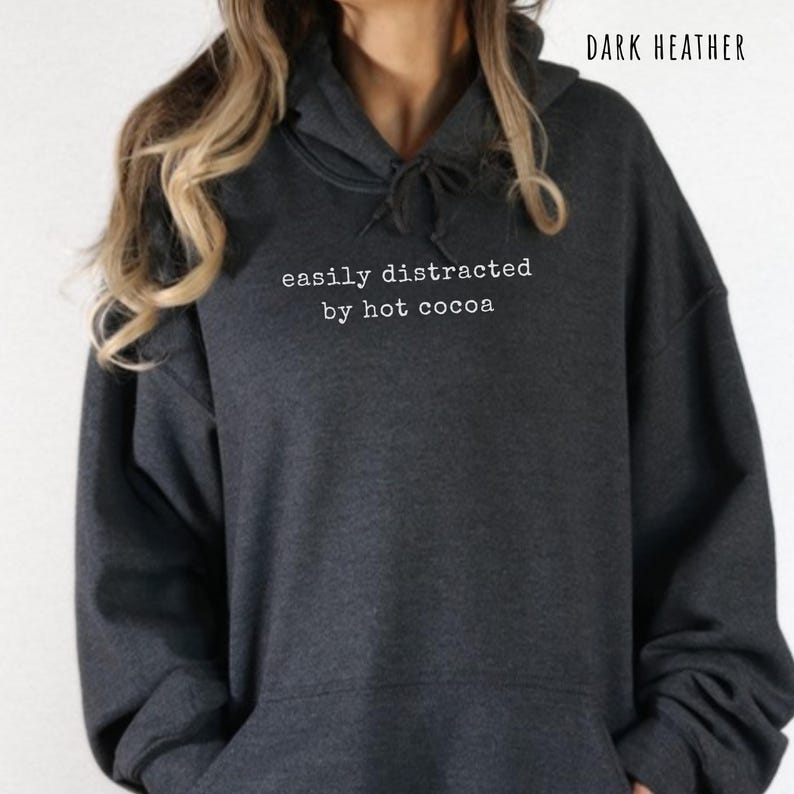 May include: A dark heather gray hooded sweatshirt featuring a drawstring hood and long sleeves. The front of the garment displays the white text "easily distracted by hot cocoa" printed in a typewriter-style font across the chest area.