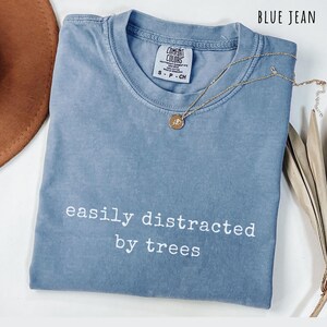 May include: A blue jean colored t-shirt with the text "easily distracted by trees" in white. A gold-colored necklace with a pendant is draped across the shirt. The shirt has a Comfort Colors tag.