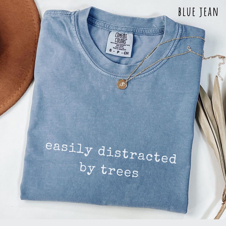 May include: A blue jean colored t-shirt with the text "easily distracted by trees" in white. A gold-colored necklace with a pendant is draped across the shirt. The shirt has a Comfort Colors tag.