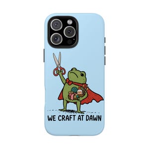 May include: A light blue phone case featuring a green frog wearing a red cape, holding scissors in one hand and a basket of craft supplies in the other. The text WE CRAFT AT DAWN is printed in black at the bottom of the phone case.