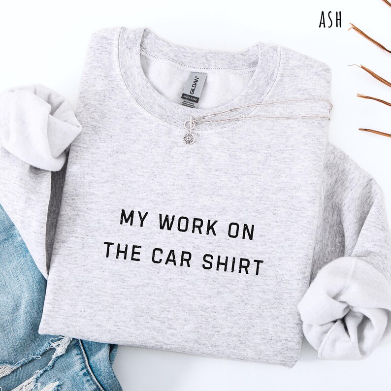 May include: Light gray sweatshirt with the text "MY WORK ON THE CAR SHIRT" in black. The sweatshirt has long sleeves and a crew neck. A silver necklace with a small pendant is draped across the front. The word "ASH" is in the top right corner.