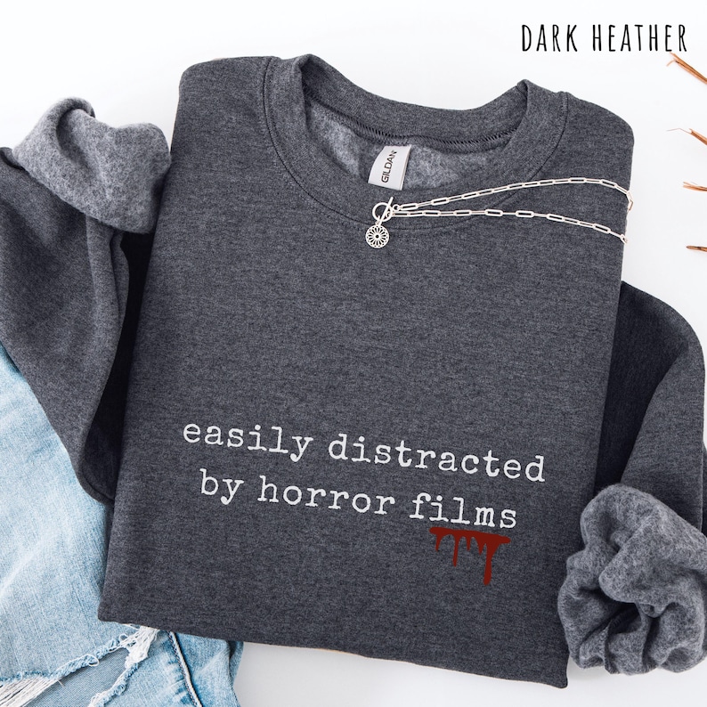 May include: Dark heather gray sweatshirt with the text "easily distracted by horror films" in white. The text is accompanied by a red blood drip graphic. A silver necklace with a sunburst pendant is draped across the top.