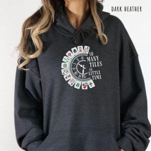 May include: Dark heather gray hoodie with a circular graphic featuring Mahjong tiles and the text "SO MANY TILES SO LITTLE TIME." The design includes a clock face and Chinese characters. The hoodie has a drawstring hood.