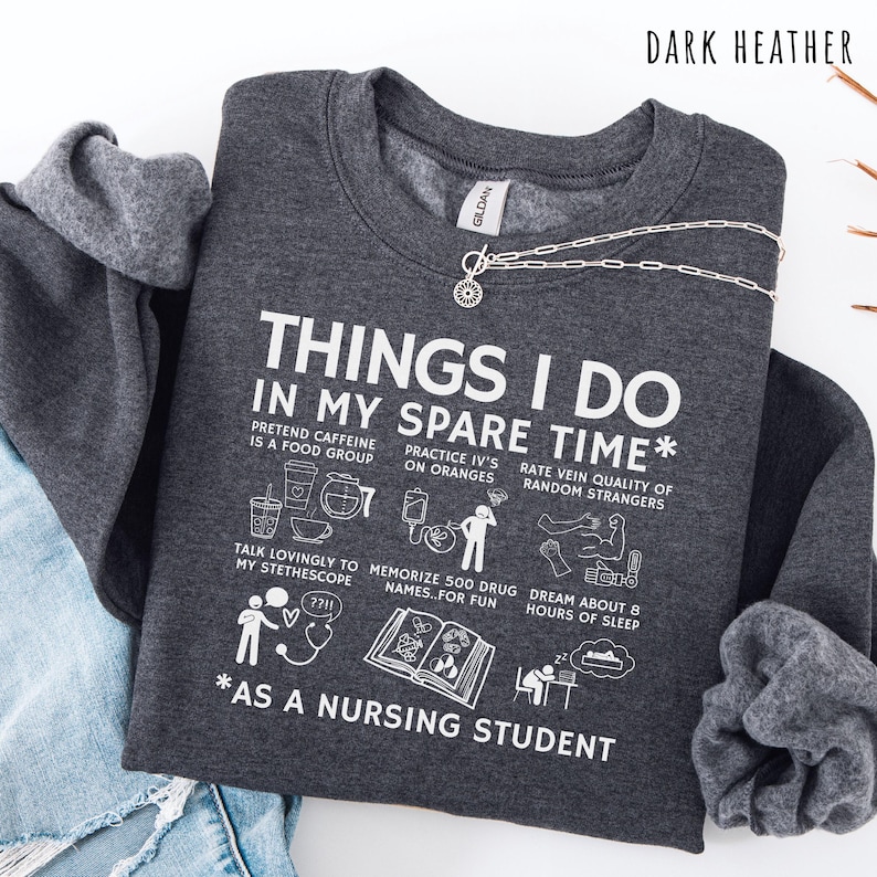 Peut inclure: Sweat-shirt gris fonc&eacute; avec texte et illustrations blanches. Le texte dit "THINGS I DO IN MY SPARE TIME *AS A NURSING STUDENT". Le design comprend des dessins de tasses &agrave; caf&eacute;, de poches IV et d'un st&eacute;thoscope. Un collier argent&eacute; avec un pendentif soleil est drap&eacute; sur le dessus.