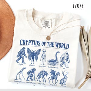 May include: An ivory t-shirt featuring a blue graphic print titled CRYPTIDS OF THE WORLD. The design displays illustrations of Bigfoot, Mothman, Grey Alien, Loch Ness Monster, Yeti, Fresno Nightcrawler, Loveland Frogman, Chupacabra, Jersey Devil, and Jackalope.