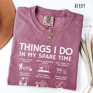 May include: Berry-colored t-shirt with the text "THINGS I DO IN MY SPARE TIME" and gardening-related icons. The shirt has a crew neck and a small tag. A gold necklace with a pendant is draped across the shirt.