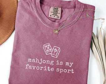May include: Berry-colored t-shirt with the text "mahjong is my favorite sport" and a graphic of mahjong tiles. A gold necklace with a pendant is draped across the shirt. The shirt is a Comfort Colors brand.