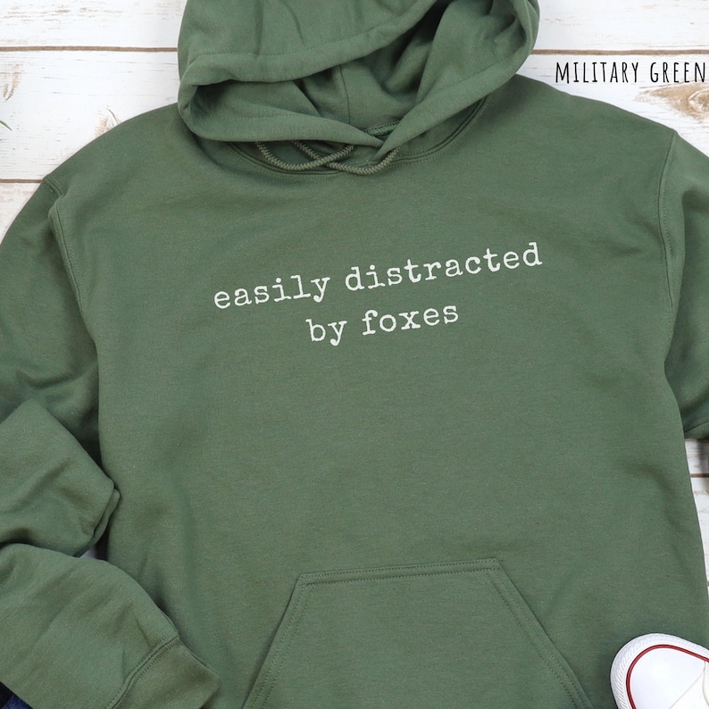 Pu&ograve; includere: Felpa con cappuccio verde militare con la scritta bianca "easily distracted by foxes". La felpa ha un cappuccio con coulisse e una tasca frontale. Il testo "MILITARY GREEN" &egrave; visibile in alto a destra.