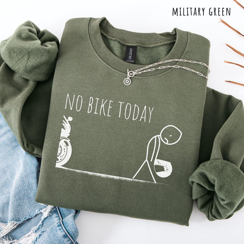 May include: Military green sweatshirt with the text "NO BIKE TODAY" and a graphic of a motorcycle and a dejected stick figure. A silver necklace with a sunburst pendant is draped on the sweatshirt. The sweatshirt is folded on top of blue jeans.