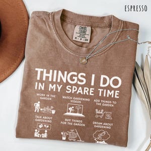 May include: Espresso-colored t-shirt with the text "THINGS I DO IN MY SPARE TIME" and gardening-related illustrations. The shirt has a crew neck and short sleeves. A silver necklace with a pendant is draped across the shirt.