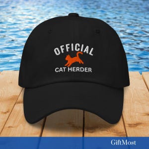 Könnte beinhalten: Schwarze Baseballkappe mit dem gestickten Text "OFFICIAL CAT HERDER" in Weiß und einer orangefarbenen Katzen-Grafik. Die Kappe liegt auf einer Holzoberfläche mit blauem Wasserhintergrund.