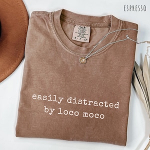 May include: Espresso-colored t-shirt with the text "easily distracted by loco moco" in white. A silver necklace with a small pendant rests on the shirt. The shirt is a Comfort Colors brand.