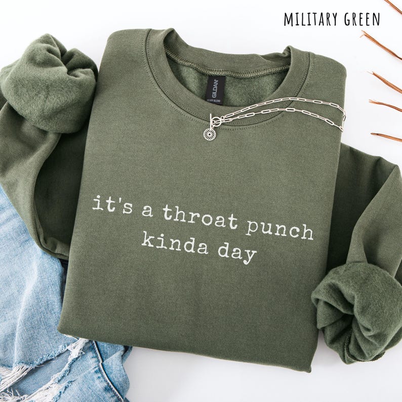 May include: Military green sweatshirt with the phrase "it's a throat punch kinda day" in white lettering. A silver necklace with a sunburst pendant rests on the sweatshirt. The sweatshirt is folded, with a portion of blue jeans visible.