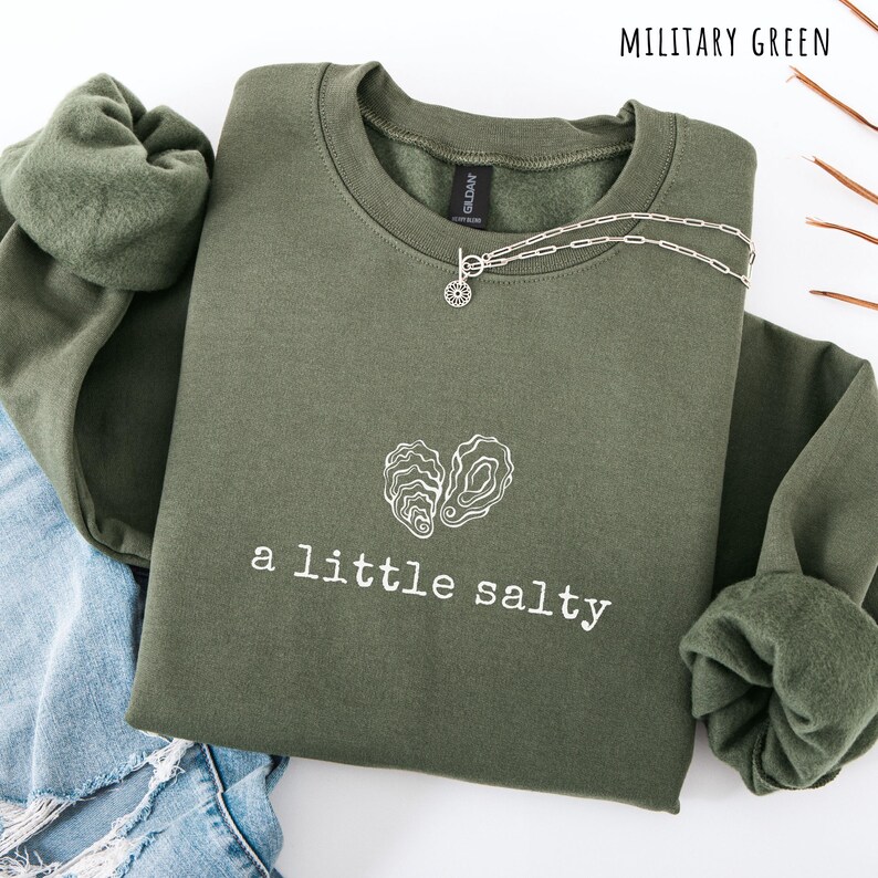 Pode incluir: Camisola verde militar com o texto "a little salty" e um gr&aacute;fico de duas conchas de ostra. Um colar de prata com um pingente de sol est&aacute; sobre a camisola. A camisola est&aacute; dobrada sobre ganga azul.