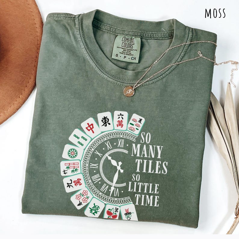 May include: Moss green t-shirt with a graphic featuring Mahjong tiles arranged around a clock face. The text reads "So Many Tiles, So Little Time." A gold necklace with a pendant rests on the shirt.