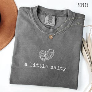 Pode incluir: Camiseta cinza-pimenta com o texto "a little salty" e um gr&aacute;fico de ostra branca. A camisa tem gola redonda e mangas curtas. Um colar de prata com um pequeno pingente est&aacute; sobre a camisa.