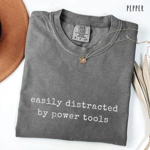 May include: Pepper-colored Comfort Colors t-shirt with the text "easily distracted by power tools" in white. A silver necklace with a small pendant is draped across the shirt. The shirt is folded and has a tag at the neckline.