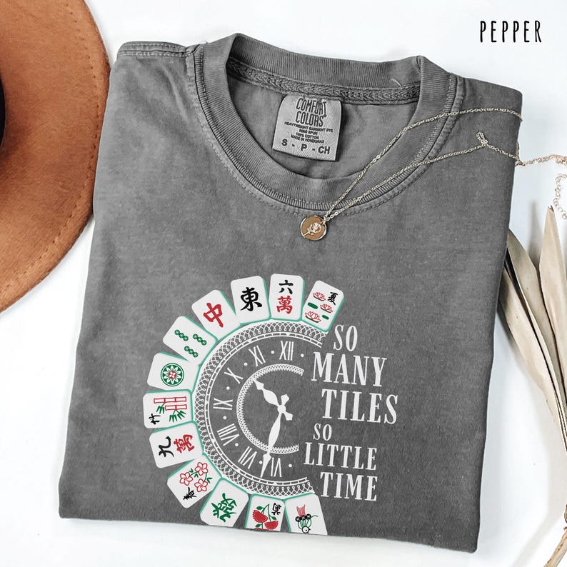 May include: A gray Comfort Colors t-shirt with a round neck and short sleeves. The shirt features a clock design with Mahjong tiles and the text "SO MANY TILES SO LITTLE TIME". A silver necklace with a pendant is on the shirt.