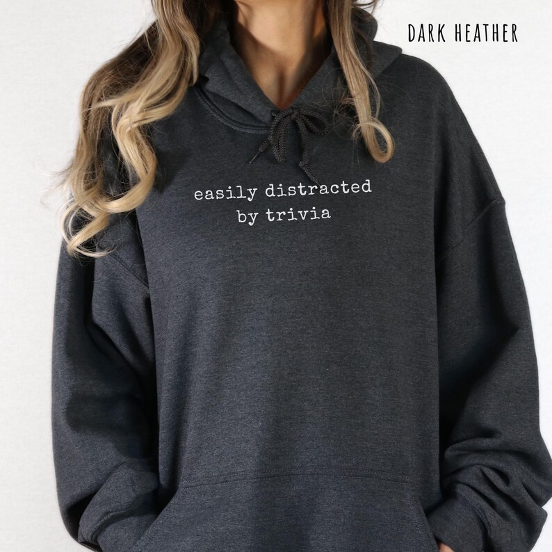 May include: Dark heather gray hoodie with the text "easily distracted by trivia" in white. The hoodie has a drawstring hood and a front pocket. The words "Dark Heather" are printed at the top right.