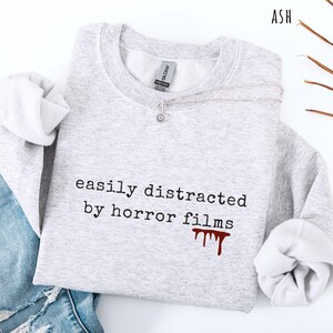 May include: Ash-colored sweatshirt with the phrase "easily distracted by horror films" printed in black. A blood-red drip graphic is below the text. A silver necklace with a small pendant rests on the sweatshirt.