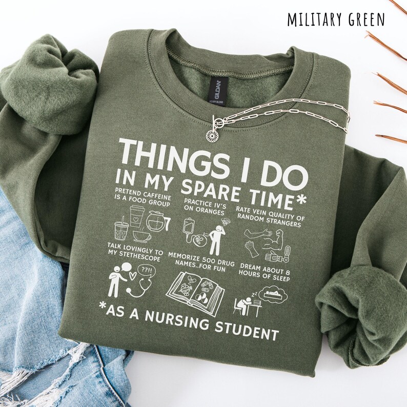 Peut inclure: Sweat-shirt vert militaire avec le texte "THINGS I DO IN MY SPARE TIME" et des illustrations. Le texte comprend des phrases comme "Pretend caffeine is a food group" et "As a nursing student". Un collier argent&eacute; est drap&eacute; sur le sweat-shirt.