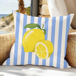 Puede incluir: Cojín cuadrado con un diseño de rayas azules y blancas. Presenta una ilustración de limón amarillo con una hoja verde. El cojín está sobre una silla de mimbre cerca de una ventana con vistas al mar.