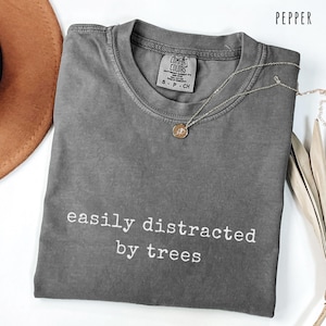May include: Pepper-colored t-shirt with the text "easily distracted by trees" in white. The shirt has a crew neck and short sleeves. A silver necklace with a small pendant is draped across the shirt. A brown hat is visible in the upper left corner.