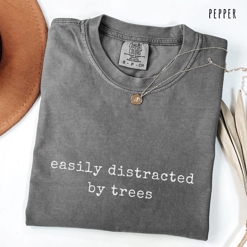 May include: Pepper-colored t-shirt with the text "easily distracted by trees" in white. The shirt has a crew neck and short sleeves. A silver necklace with a small pendant is draped across the shirt. A brown hat is visible in the upper left corner.