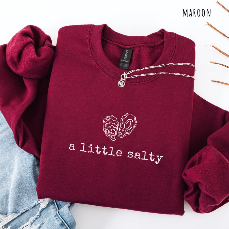 Pode incluir: Camisola bord&ocirc; com texto branco e um desenho de conchas. O texto diz "a little salty". Um colar de prata com um pingente de sol est&aacute; na camisola. A palavra "MAROON" est&aacute; no canto superior direito.