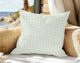 May include: A square decorative pillow with a light green and white gingham pattern. The pillow is placed on a wicker chair near a window overlooking the ocean. The pillow's pattern is a classic, timeless design.