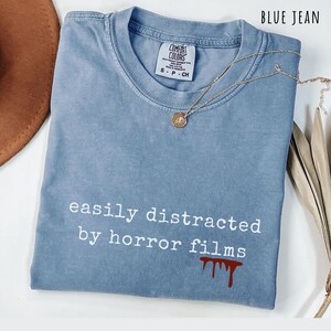 May include: Blue jean-colored Comfort Colors t-shirt with the text "easily distracted by horror films" and a blood splatter graphic. A gold necklace with a pendant rests on the shirt. The shirt is folded.