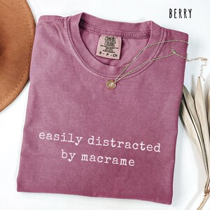 May include: Berry-colored t-shirt with the text "easily distracted by macrame" in white. A gold necklace with a pendant is on the shirt. The shirt is folded and has a Comfort Colors tag. A brown hat is visible in the background.