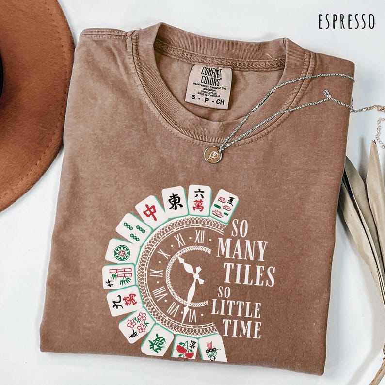 May include: Espresso-colored t-shirt with a graphic featuring Mahjong tiles and the text "SO MANY TILES SO LITTLE TIME." A silver necklace with a pendant rests on the shirt. The shirt is labeled "Comfort Colors" and is size S-CH.