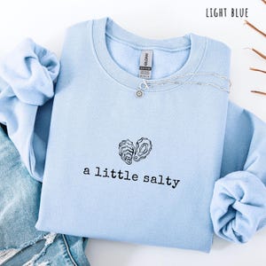 Pode incluir: Camisola azul clara com a frase "a little salty" e um desenho de duas conchas de ostra. Um colar de prata com um pingente de sol est&aacute; na camisola. As palavras "LIGHT BLUE" est&atilde;o no canto superior direito.