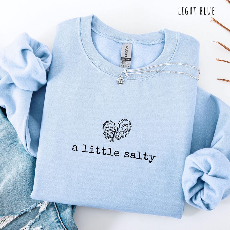 Pode incluir: Camisola azul clara com a frase "a little salty" e um desenho de duas conchas de ostra. Um colar de prata com um pingente de sol est&aacute; na camisola. As palavras "LIGHT BLUE" est&atilde;o no canto superior direito.