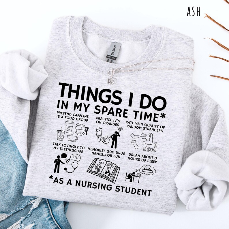 Peut inclure: Sweat-shirt gris clair avec le texte "THINGS I DO IN MY SPARE TIME *AS A NURSING STUDENT" et des illustrations humoristiques. Le sweat-shirt a un col rond et des manches longues. Un collier argent&eacute; avec un petit pendentif est &eacute;galement visible.