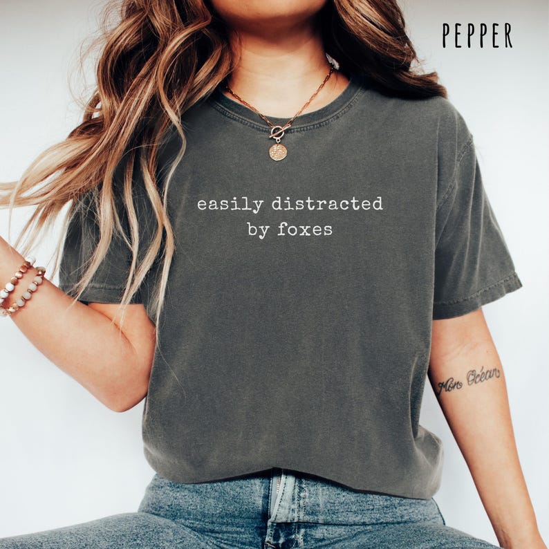 Pu&ograve; includere: T-shirt grigio scuro con la scritta bianca "easily distracted by foxes". La maglietta &egrave; a girocollo e sembra essere realizzata in un materiale morbido. La parola "PEPPER" &egrave; stampata nell'angolo in alto a destra.