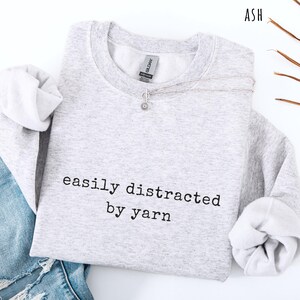 May include: A light gray sweatshirt with the text "easily distracted by yarn" in black. A silver necklace with a small pendant rests on the sweatshirt. The sleeves are cuffed, and a portion of blue jeans is visible.