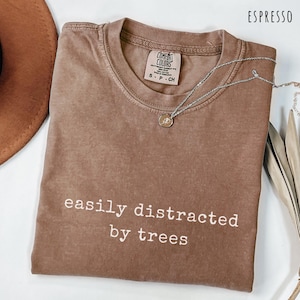 May include: Espresso-colored t-shirt with the text "easily distracted by trees" in white. The shirt has a crew neck and short sleeves. A silver necklace with a pendant is draped across the shirt.