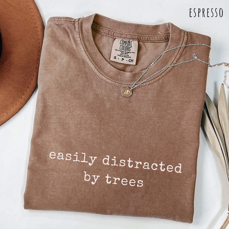May include: Espresso-colored t-shirt with the text "easily distracted by trees" in white. The shirt has a crew neck and short sleeves. A silver necklace with a pendant is draped across the shirt.