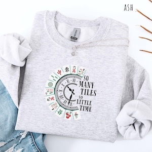 May include: A light gray sweatshirt with a circular graphic featuring a clock design and Mahjong tiles. The text "SO MANY TILES SO LITTLE TIME" is printed on the right side of the clock. A silver necklace with a small pendant is draped across the sweatshirt.