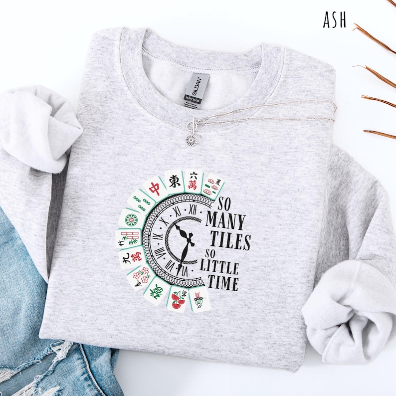 May include: A light gray sweatshirt with a circular graphic featuring a clock design and Mahjong tiles. The text "SO MANY TILES SO LITTLE TIME" is printed on the right side of the clock. A silver necklace with a small pendant is draped across the sweatshirt.
