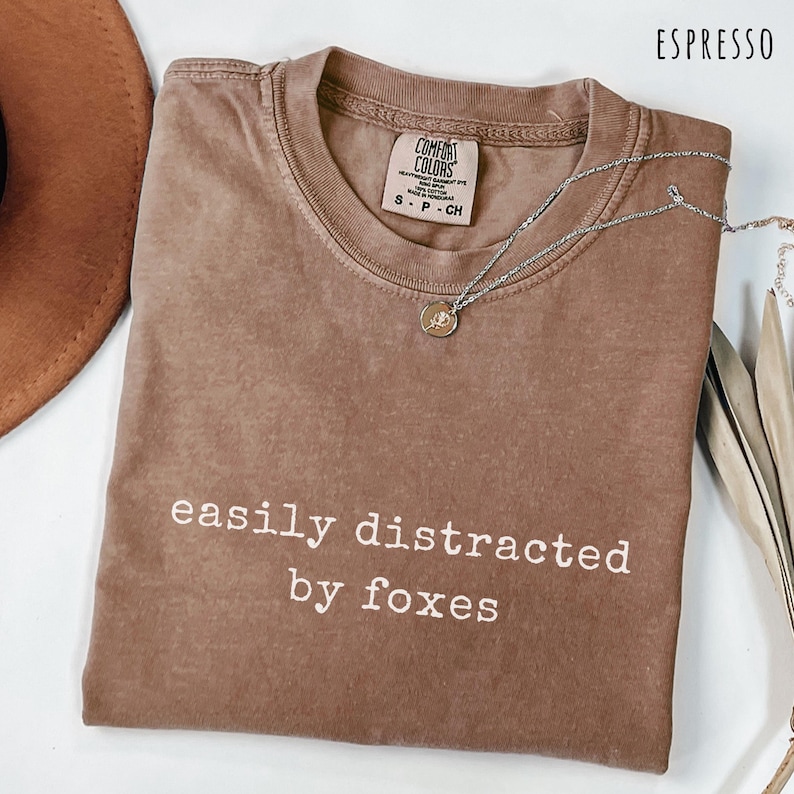 Brown shirt with the phrase “easily distracted by foxes” folded neatly beside a hat and necklace, a cute funny fox lover tee for nature and wildlife fans.