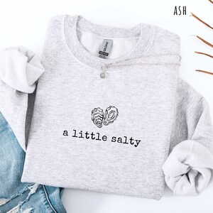 Pode incluir: Camisola cinza claro com a frase "a little salty" e um desenho em forma de cora&ccedil;&atilde;o feito com conchas de ostra. Um colar de prata com um pequeno pendente est&aacute; sobre a pe&ccedil;a de roupa. A palavra "ASH" &eacute; vis&iacute;vel no canto superior direito.
